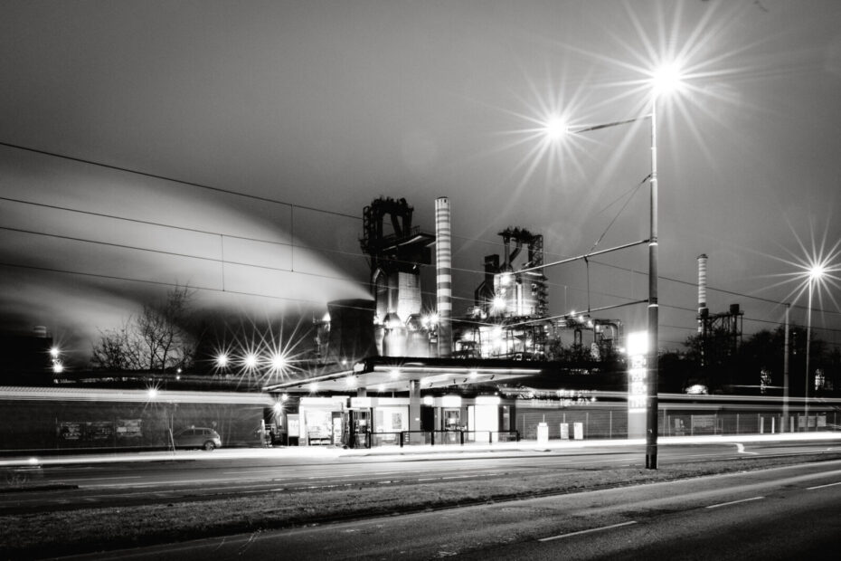 Hochofenwerk hinter eine Tankstelle, im Vordergrundrund die Hauptstraße mit Straßenbahnschienen und Oberleitung