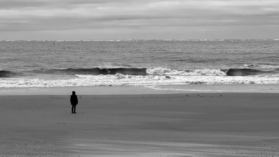 Schwarz-weiß Bild mit hohen Kontrast, ein Mensch an einem ansonsten leeren Strand, den Blick aufs Meer gerichtet, wo eine Welle gerade zu brechen beginnt