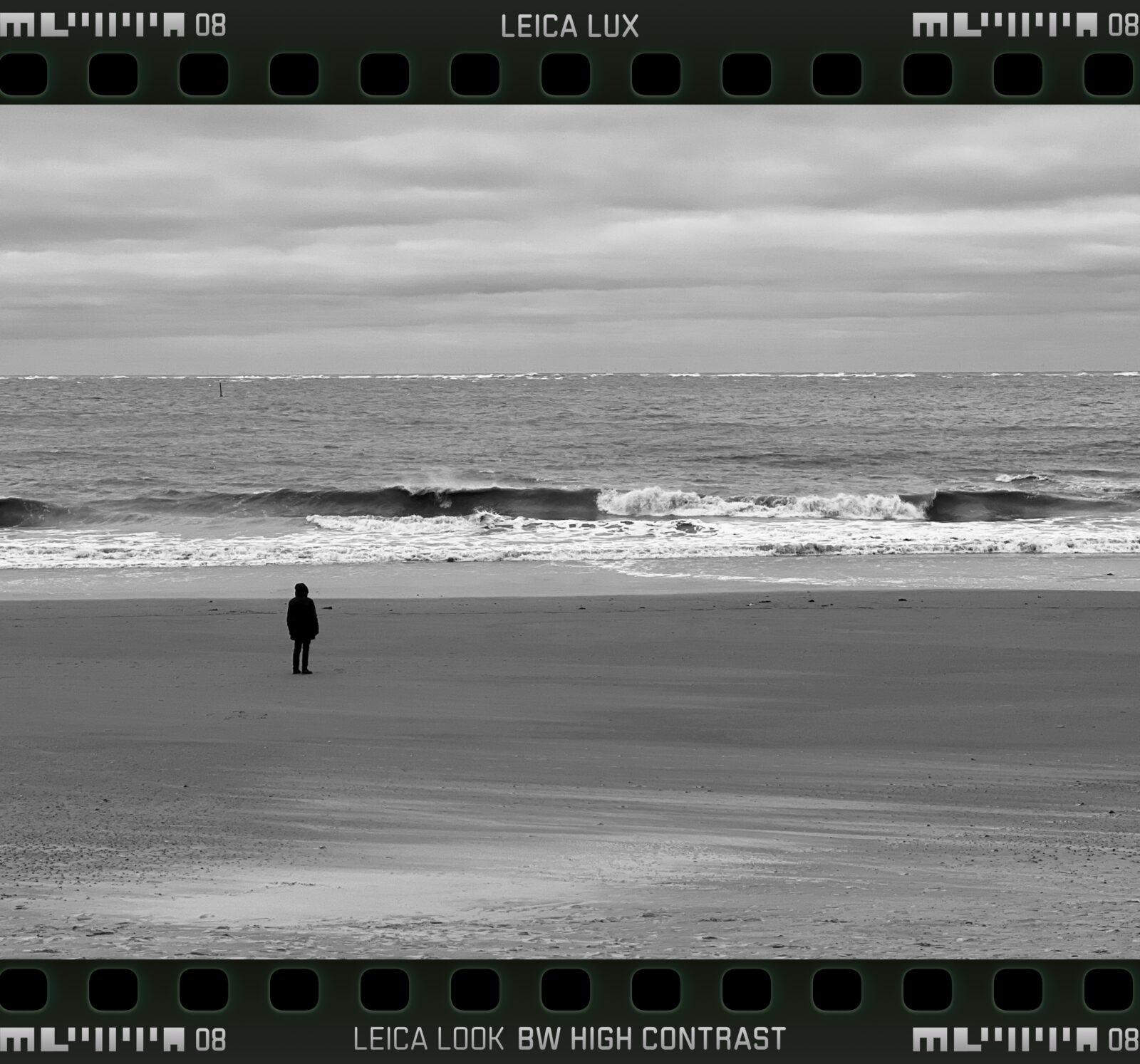 Schwarz-weiß Bild mit hohen Kontrast, ein Mensch an einem ansonsten leeren Strand, den Blick aufs Meer gerichtet, wo eine Welle gerade zu brechen beginnt