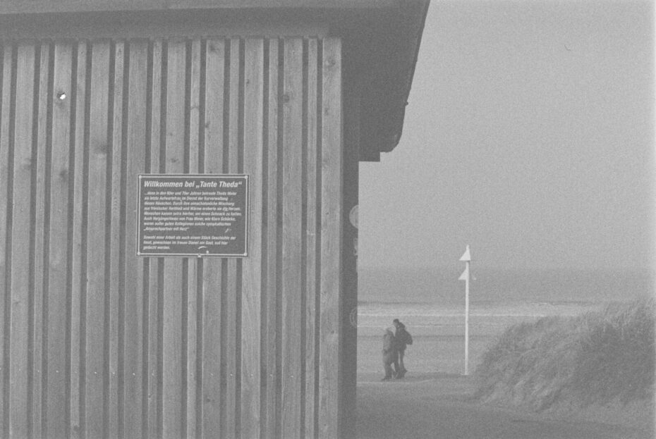 Schwarz- Weiß Bild mit grobem Korn, Links eine Fassade, daneben ein Durchgang und Menschen am Strand