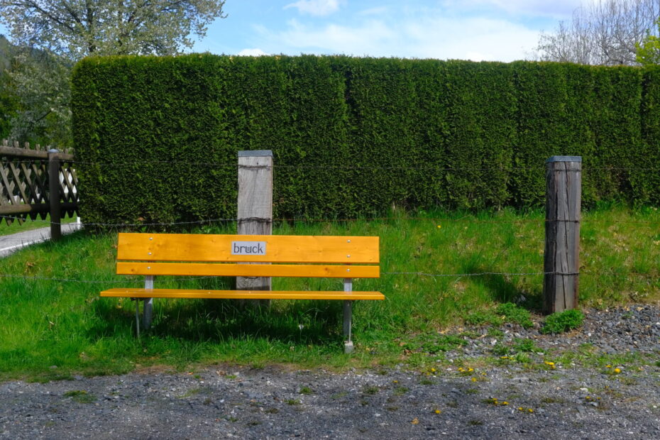 Bank vor Stacheldraht und grüner Hecke mit der Aufschrift "Bruck"
