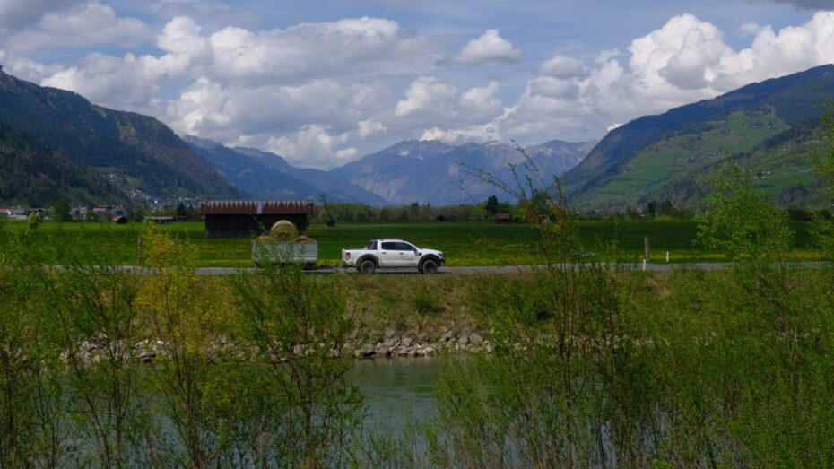 Ein Pickup fährt mit einem Anhänger mit Heuballen an der Salzach entlang, im Hintergrund die Berge
