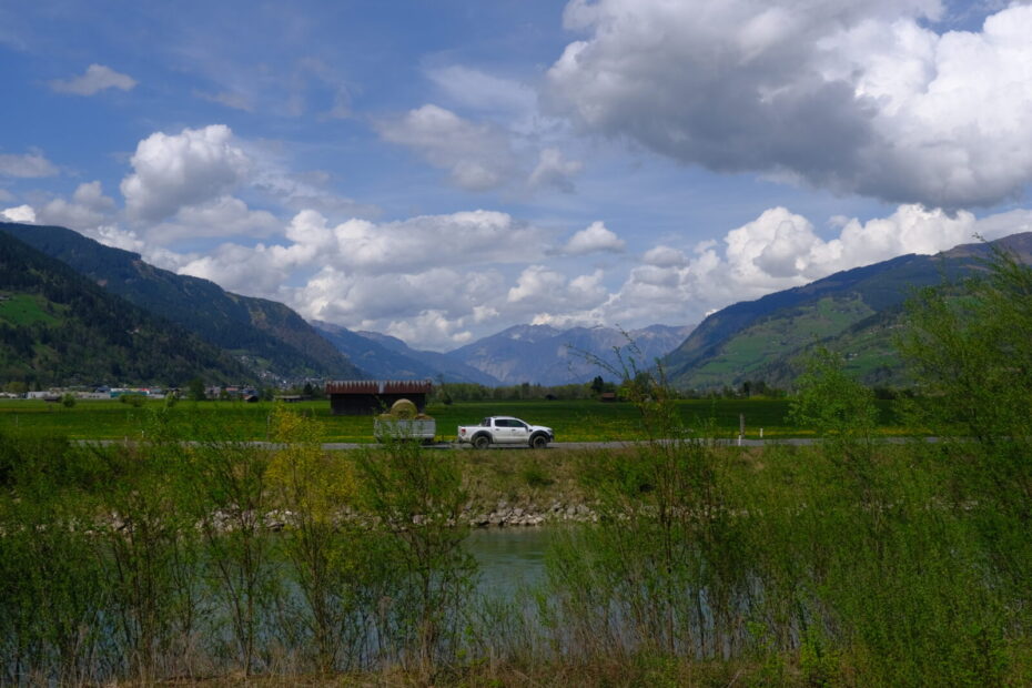 Ein Pickup fährt mit einem Anhänger mit Heuballen an der Salzach entlang, im Hintergrund die Berge