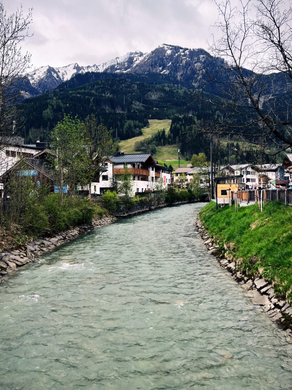 Blick auf die Kapruner Ache, eine Fluss, der von den Bergen durch Kaprun führt, Häuser säumen das Ufer, der Fluss macht eine Biegung nach rechts, im Hintergrund die Berge rund um das Kitzsteinhorn