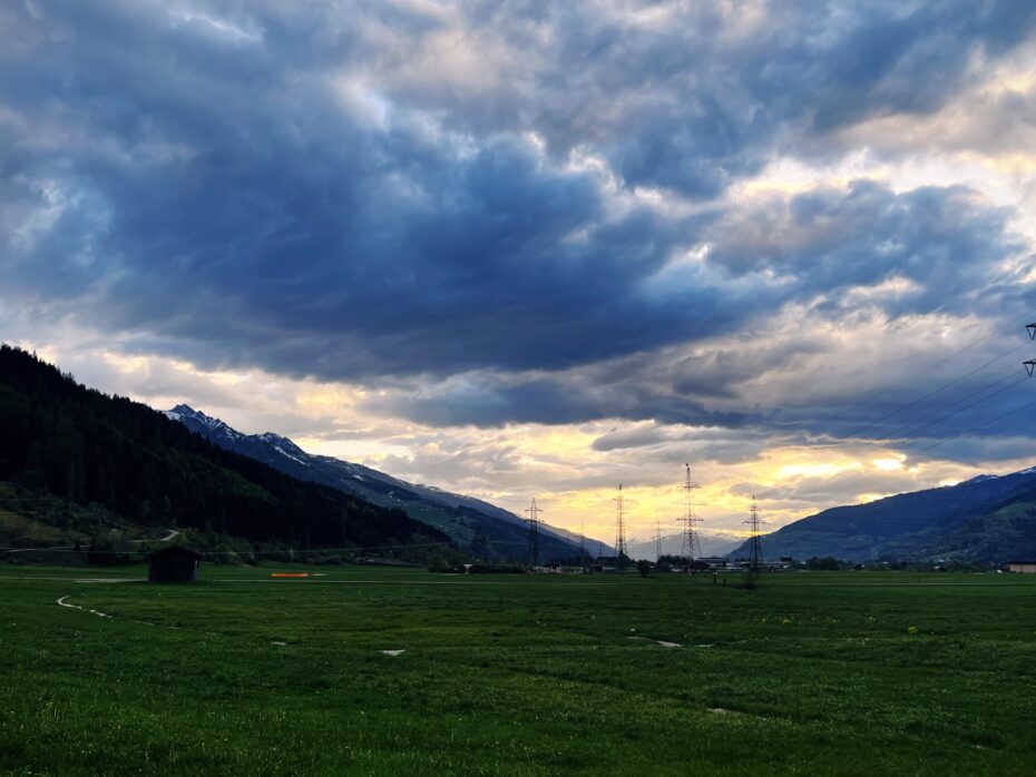 Blick auf eine weite Talfläche, eingerahmt von Bergen, die Sonne geht unter, die Wolken bilden ein Dramatik