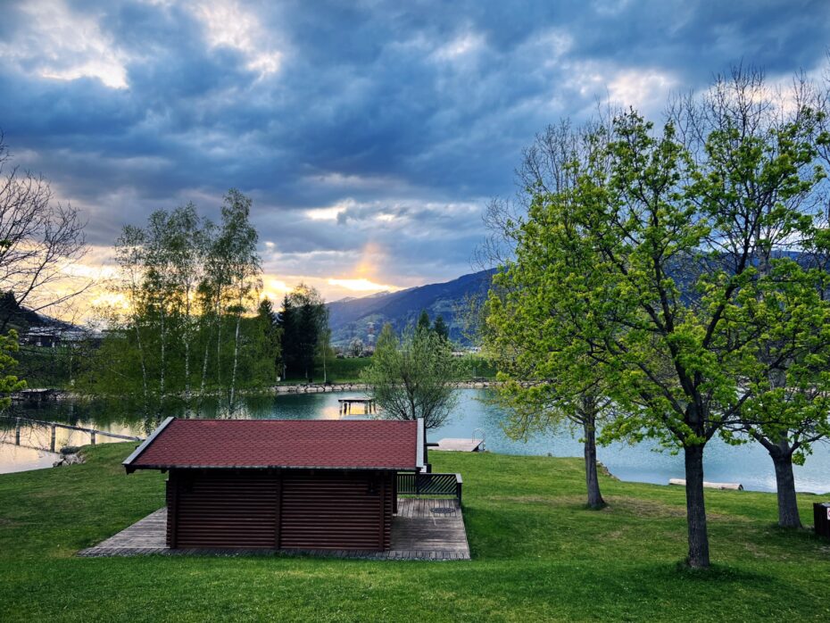 Blick auf einen Badesee in Bruck / Glocknerstr., im Hintergrund der Sonnenuntergang hinter den Bergen, Himmel bewölkt, im Vordergrund ein kleines Holzhaus