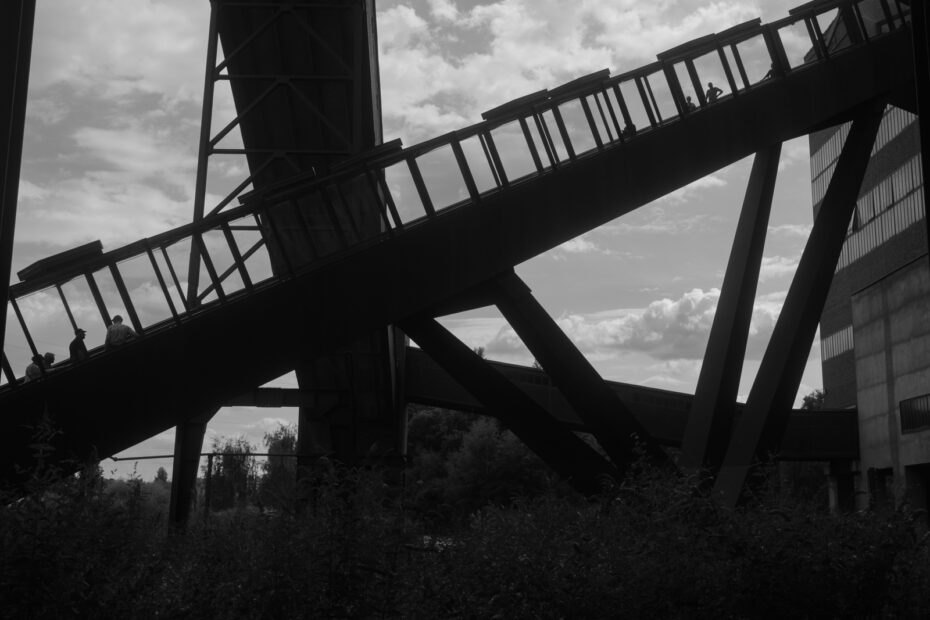 Schwarz-Weiß Bild von der Seite der Rolltreppe, die zum Ruhrmuseum auf dem Geländer der Zeche Zollverein führt, auf der Menschen nach oben fahren oder von oben kommen
