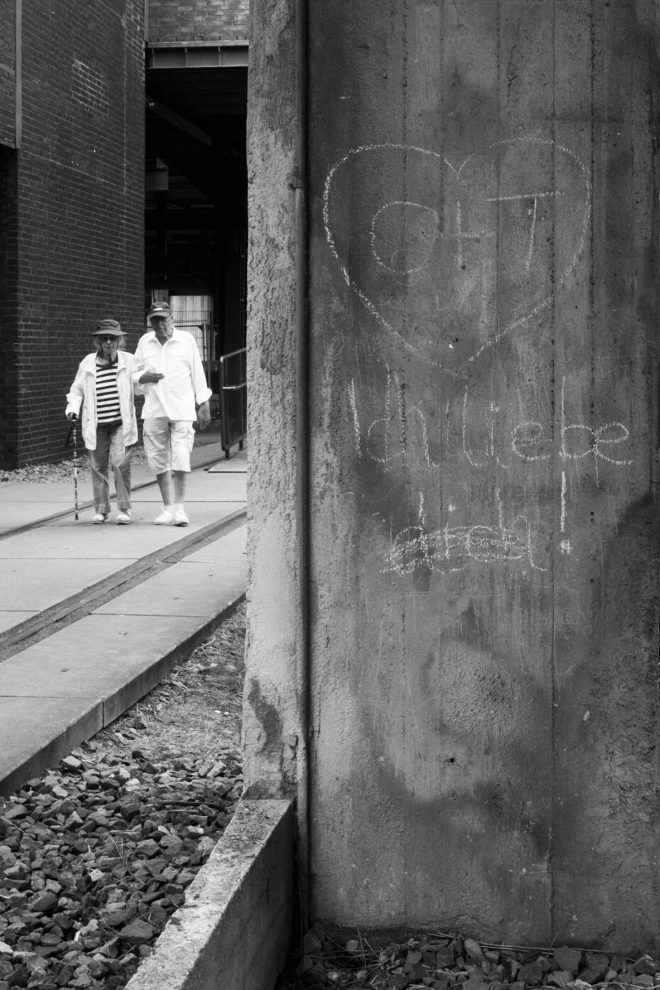 Schwarz-Weiß Bild, auf dem auf einem Betonpfeiler ein Herz mit Inschrift "O+T" gemalt ist und mit "Ich liebe dich!" unterschrieben ist. Links davon läuft ein älteres Paar eingehakt in die Szenerie