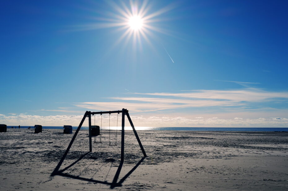 Eine Schaukel steht in der vollen Herbssonne am Strand, der tiefere Sonnenstand lässt Schatten erwachsen, im Hintergrund Strandkörbe
